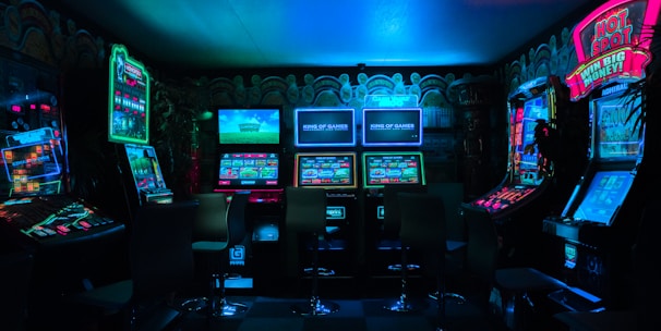 Dimly lit gaming arcade featuring a variety of brightly illuminated slot machines and video gaming screens. Neon signs and colorful displays create a vibrant atmosphere. The arrangement of gaming machines suggests an inviting space for entertainment, with chairs positioned for players.