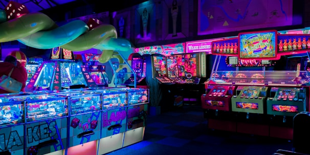 An indoor arcade filled with vibrant gaming machines lit with neon blue and pink lights. The scene includes various arcade games with flashing screens and colorful displays, and a large overhead structure resembling a green snake. The background shows illuminated signs and themed decor, contributing to an immersive entertainment atmosphere.