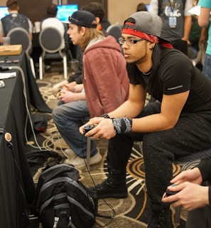 Several people are sitting in a room with gaming consoles and monitors, focusing intently on their screens. The setting appears to be a casual gaming event, with participants holding controllers while playing video games. The room has a casual atmosphere with chairs and tables set up for gaming.