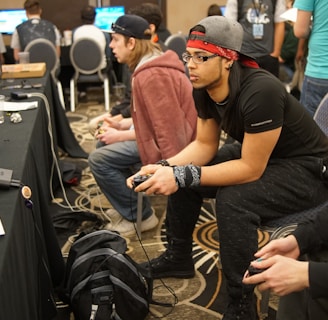 Several people are sitting in a room with gaming consoles and monitors, focusing intently on their screens. The setting appears to be a casual gaming event, with participants holding controllers while playing video games. The room has a casual atmosphere with chairs and tables set up for gaming.