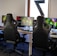 A row of gaming stations equipped with large monitors, gaming chairs, keyboards, and headphones. The setup is organized and ready for gaming, featuring a colorful screen display. The room has a modern and tech-savvy ambiance.