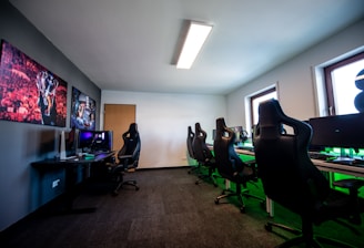 A well-lit gaming room features multiple gaming stations with high-backed chairs and monitors. The room has a minimalist setup with green and black color tones. Large posters adorn the walls, and the room appears clean and organized.
