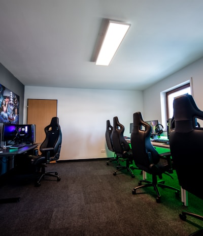 A well-lit gaming room features multiple gaming stations with high-backed chairs and monitors. The room has a minimalist setup with green and black color tones. Large posters adorn the walls, and the room appears clean and organized.