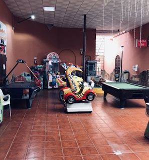 An indoor arcade area featuring various gaming machines, including an air hockey table and a ride-on toy car. The room has a tiled floor and several colorful gaming devices scattered around. Chairs and arcade signage are visible, creating a playful and entertainment-focused atmosphere.