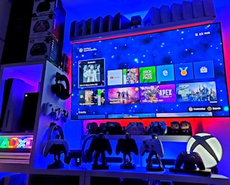 A modern gaming setup featuring a large wall-mounted screen displaying a gaming console interface. The room is decorated with gaming-themed items such as controllers and collectibles. The ambient lighting includes shades of blue and red, creating a vibrant atmosphere.