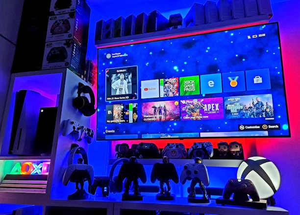 A modern gaming setup featuring a large wall-mounted screen displaying a gaming console interface. The room is decorated with gaming-themed items such as controllers and collectibles. The ambient lighting includes shades of blue and red, creating a vibrant atmosphere.