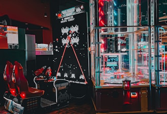 An arcade setting featuring a classic Space Invaders game setup and a modern gaming machine with seats. Bright neon lights illuminate the area, with scores displayed on digital screens. A bright and visually engaging environment full of electronic components.