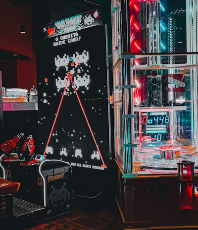 An arcade setting featuring a classic Space Invaders game setup and a modern gaming machine with seats. Bright neon lights illuminate the area, with scores displayed on digital screens. A bright and visually engaging environment full of electronic components.