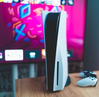 A gaming console is placed on a wooden table with a game controller nearby. In the background, a television screen displays colorful graphics, including geometric shapes and vibrant colors.