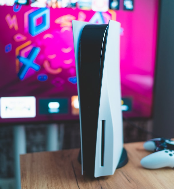 A gaming console is placed on a wooden table with a game controller nearby. In the background, a television screen displays colorful graphics, including geometric shapes and vibrant colors.