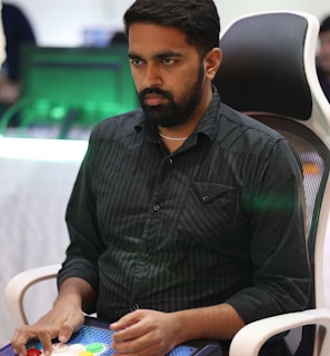 A person is seated in a chair, focused intently on something, with hands positioned on a gaming console or arcade controller. The setup appears to be indoors, with visible screens and lighting in the background, suggesting an electronic gaming environment.