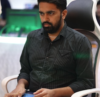 A person is seated in a chair, focused intently on something, with hands positioned on a gaming console or arcade controller. The setup appears to be indoors, with visible screens and lighting in the background, suggesting an electronic gaming environment.